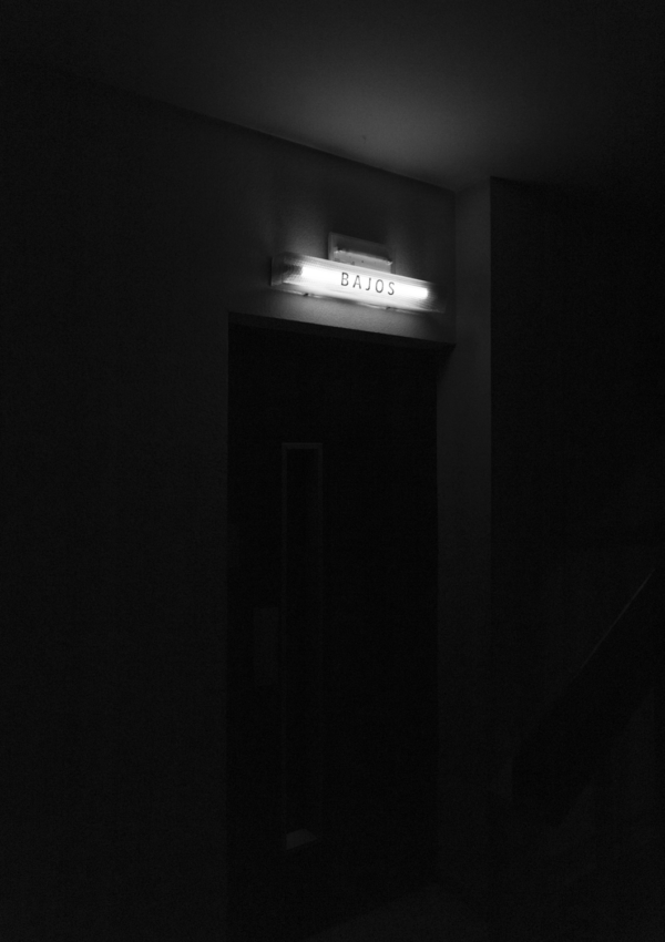Rellano de edificio en Barcelona con cartel luminoso "Bajos". Fotografía blanco y negro